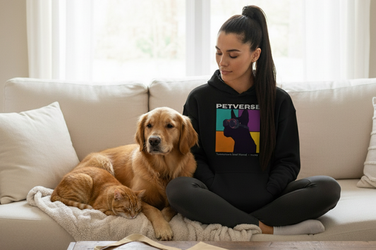 A woman in a black hoodie and dark pants sits cross-legged on a couch, holding a white mug. A golden retriever lies next to her, with an orange cat sleeping by its side. A tote bag is on the coffee table in front of them.